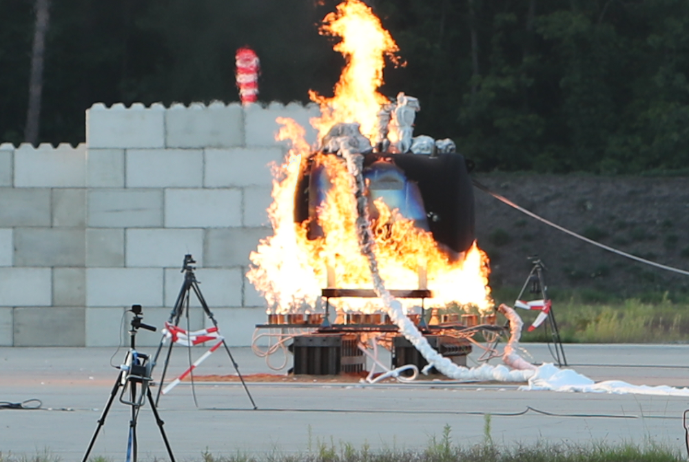 Au&szlig;enversuch mit kontrolliertem Brand: Ein gro&szlig;er zylindrischer Beh&auml;lter steht in Flammen, umgeben von Messaufbauten und Kamerastativen auf einem abgesperrten Testgel&auml;nde.