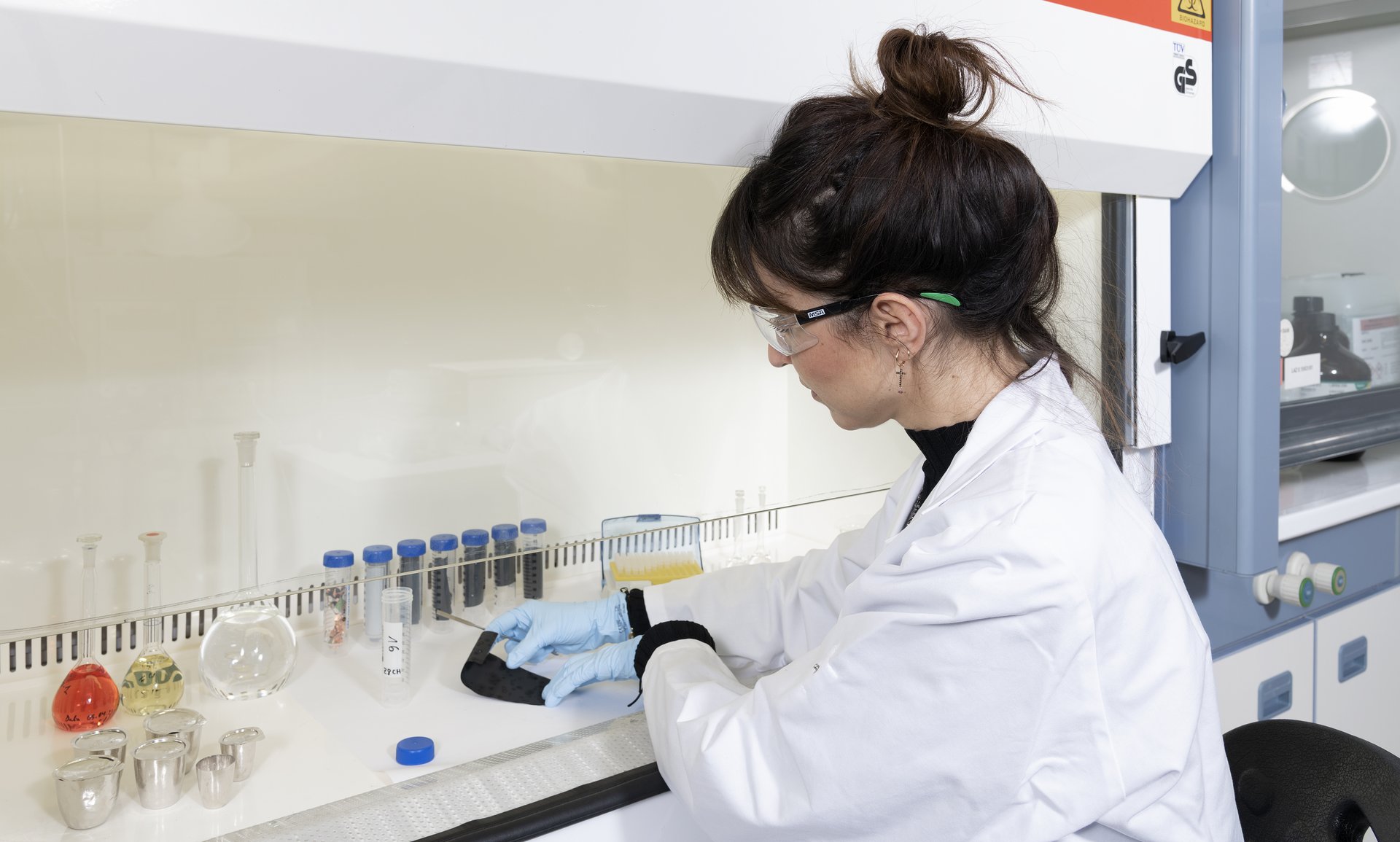Lithium Batterien Alterserkennung Dalia Morcillo sitzend im Labor an einem Tisch, von schräg hinten zu sehen, bei der Probenvorbereitung und Extraktion des Aktivmaterials von der Elektrode. Sie trägt einen Laborkittel, eine Schutzbrille und Handschuhe.