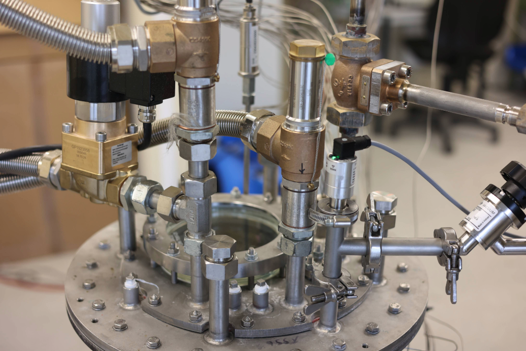 Nahaufnahme einer technischen Versuchsanlage mit Metallrohren, Ventilen, Sensoren und Verschraubungen auf einem runden Behälterdeckel in einer Laborumgebung.